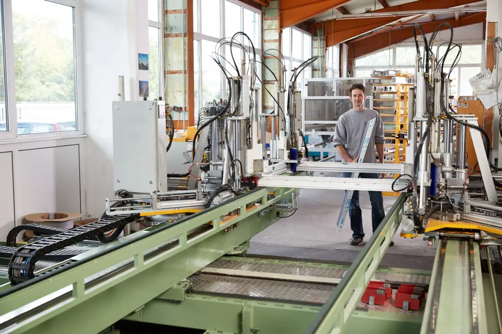 Junger Mann steht in einer Werkhalle neben einer großen grünen Industriemaschine mit Metallrahmen und Steuerungselementen