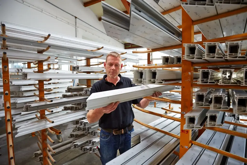 Mann in schwarzem Poloshirt hält ein langes Aluminiumprofil und prüft das Material in einem Lager mit vielen weiteren Aluminiumprofilen auf Regalen