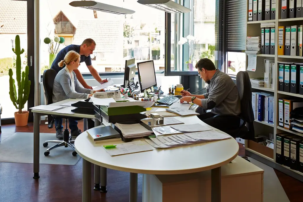 Drei Personen arbeiten an Schreibtischen in einem Büro mit großen Fenstern und Regalen voller Ordner
