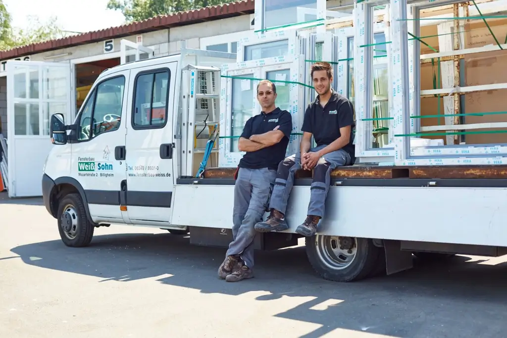 Zwei Männer in Arbeitskleidung lehnen an einem weißen Transporter mit Fensterelementen auf der Ladefläche