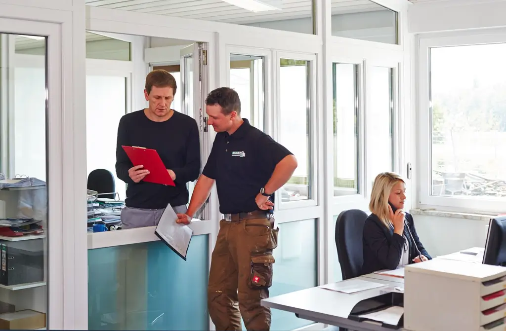 Zwei Männer stehen in einem Büro und besprechen sich während eine Frau am Schreibtisch telefoniert