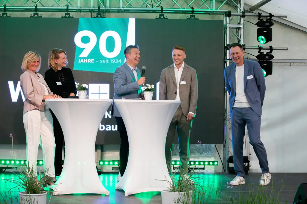 Fünf Personen stehen lächelnd an zwei Stehtischen auf einer Bühne vor einer großen Leinwand mit der Aufschrift '90 Jahre seit 1934'