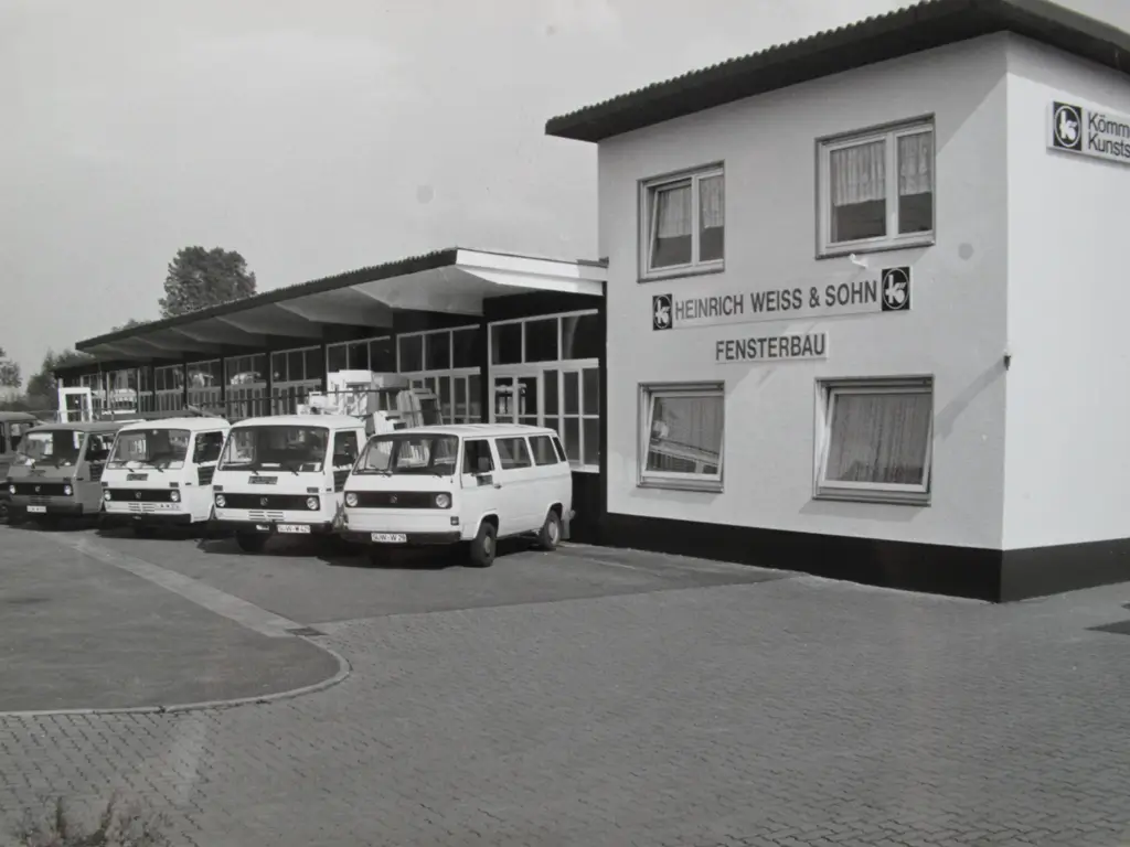 Schwarz-weiße Aufnahme eines Firmengebäudes mit mehreren geparkten Kleintransportern und der Aufschrift 'Heinrich Weiss & Sohn Fensterbau'