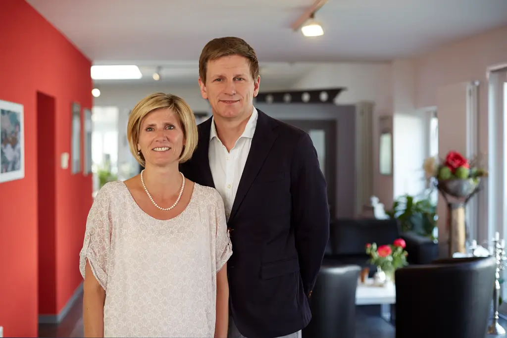 Zwei Personen, eine Frau mit kurzen blonden Haaren und Perlenkette und ein Mann mit kurzem braunem Haar, stehen nebeneinander in einem hellen Flur mit roten und weißen Wänden.