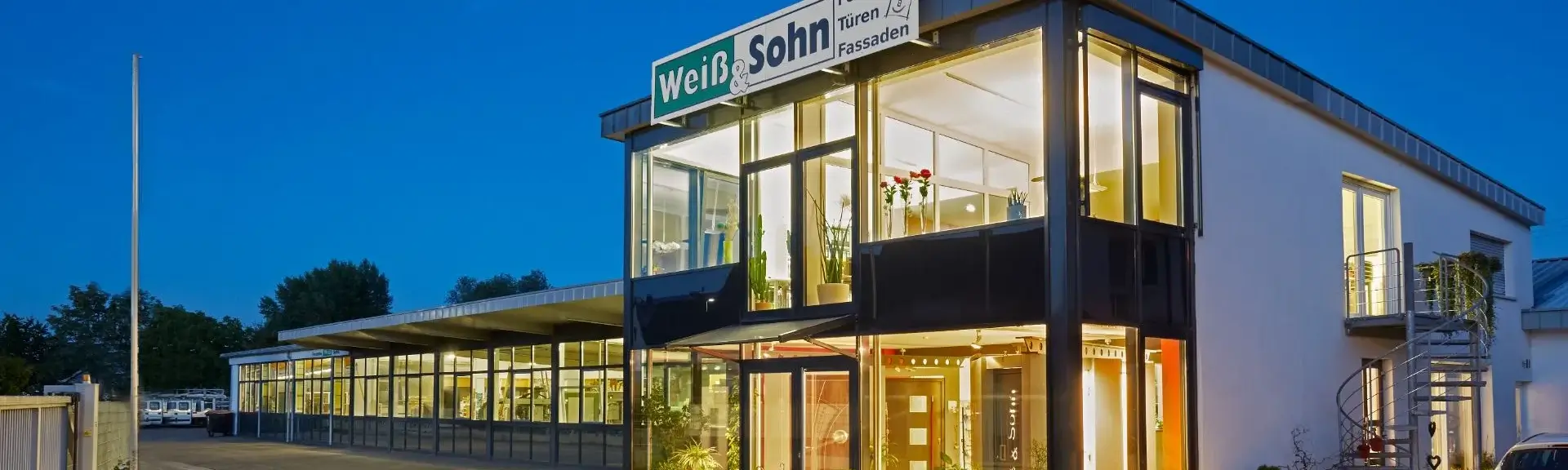 Weiß-Sohn-Glaserei-Firmengebaeude Beleuchtetes Bürogebäude mit Glasfassade und Schild 'Weiß Sohn Fenster Türen Fassaden' bei Abendlicht